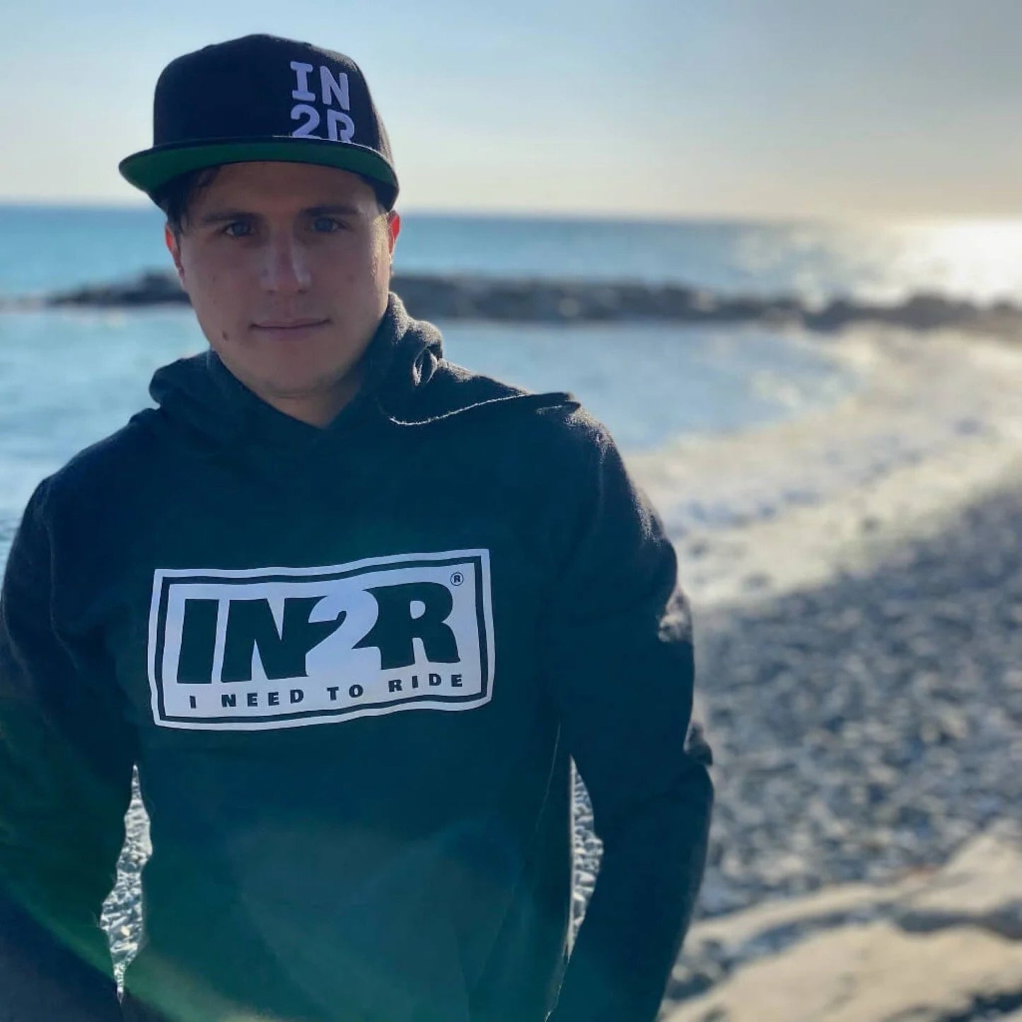 A man wearing a black and white IN2R baseball cap and a black hoodie with a large white and green IN2R logo on the chest, standing on a beach with the ocean in the background.