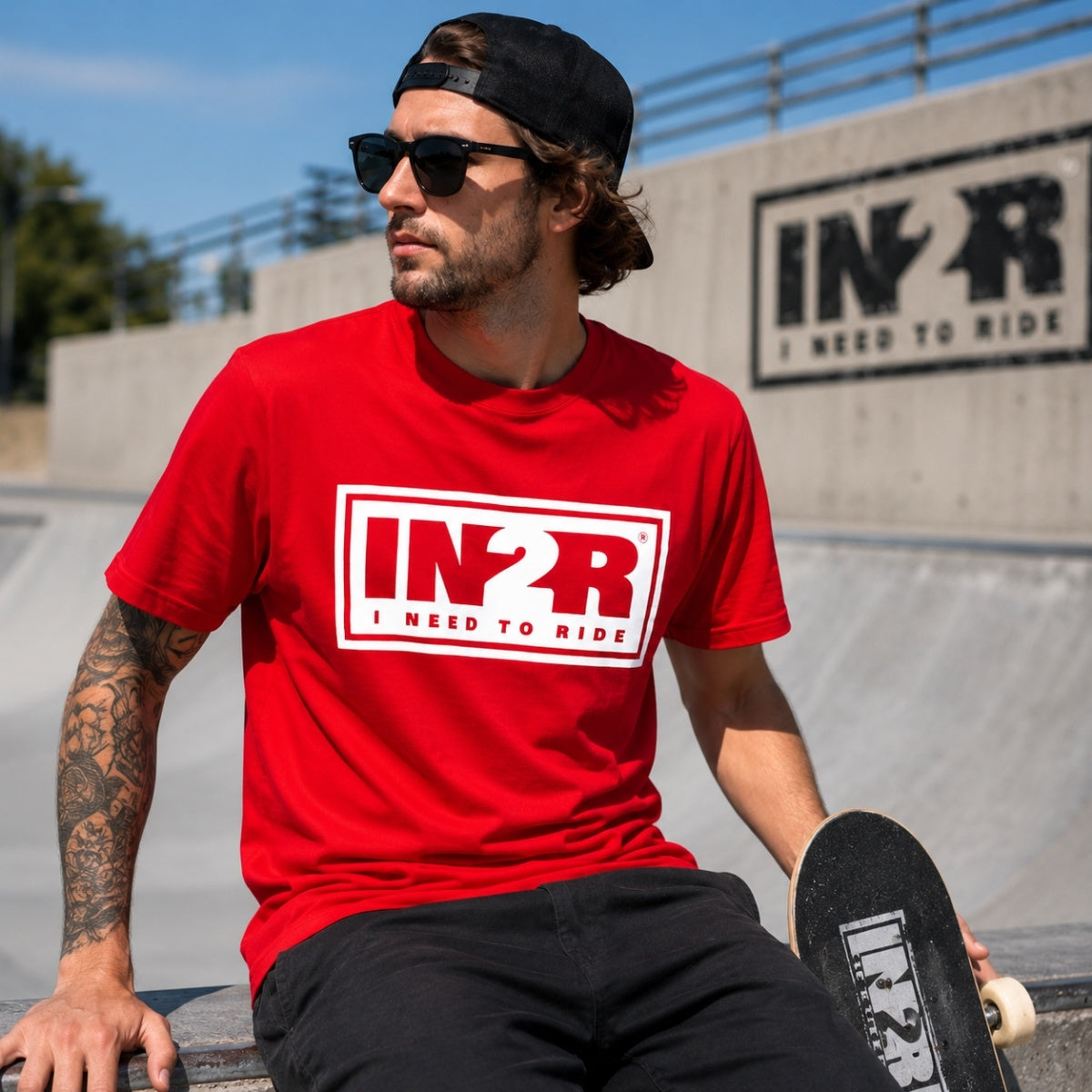 A man wearing a bright red t-shirt with a white and black 'IN2R I NEED TO RIDE' graphic, black pants, and a black cap, sitting at a skatepark with a skateboard.
