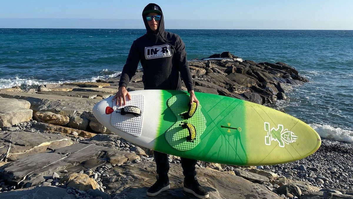 Marco, IN2R ambassador, holding a kiteboard on a rocky coastline wearing a black IN2R Original hoodie.