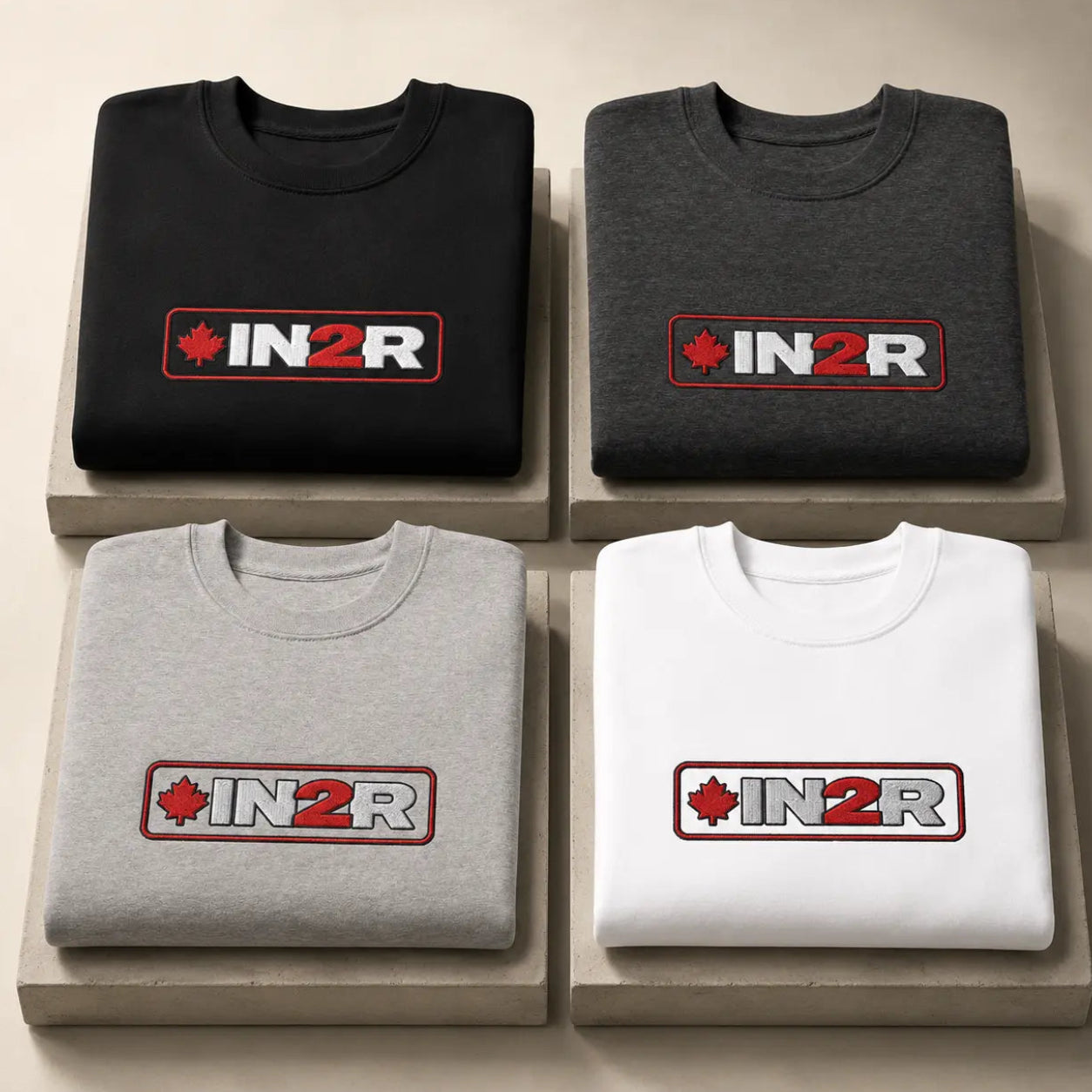Four folded crewneck sweatshirts arranged in a 2x2 grid on light beige display blocks. The top-left is black, the top-right is dark gray, the bottom-left is light gray, and the bottom-right is white.
