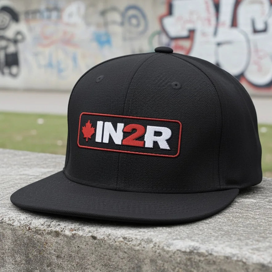 IN2R Canadian Edition black flat brim snapback hat with embroidered red maple leaf and white IN2R logo featuring a red 2, photographed on concrete against an urban graffiti background.