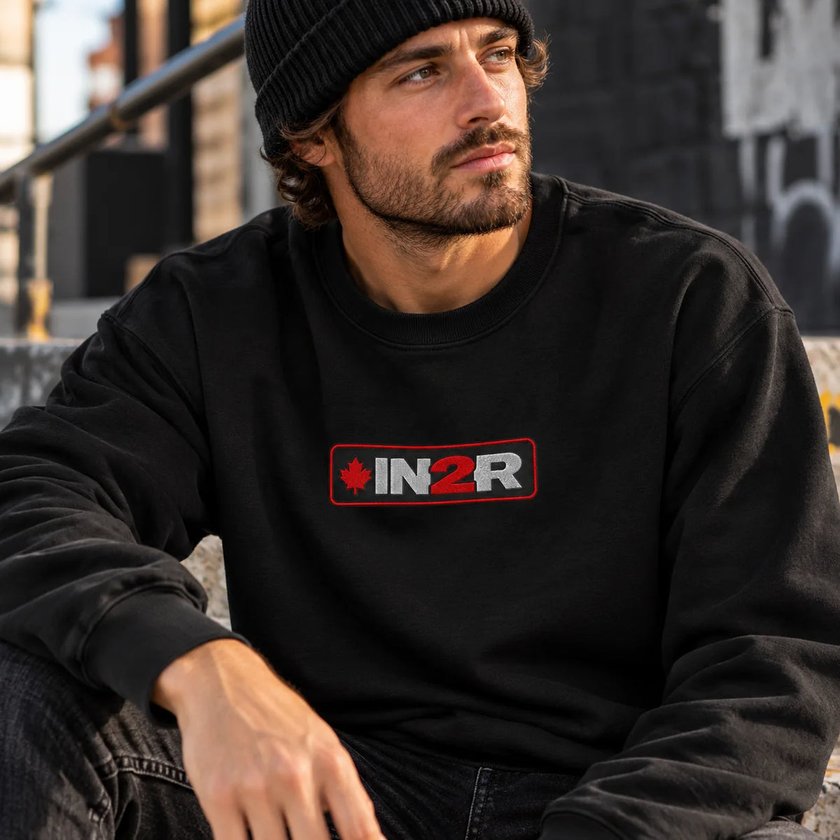 Man wearing a black IN2R Canadian Edition crewneck sweatshirt with red and white embroidered maple leaf logo, seated on concrete steps in an urban setting.