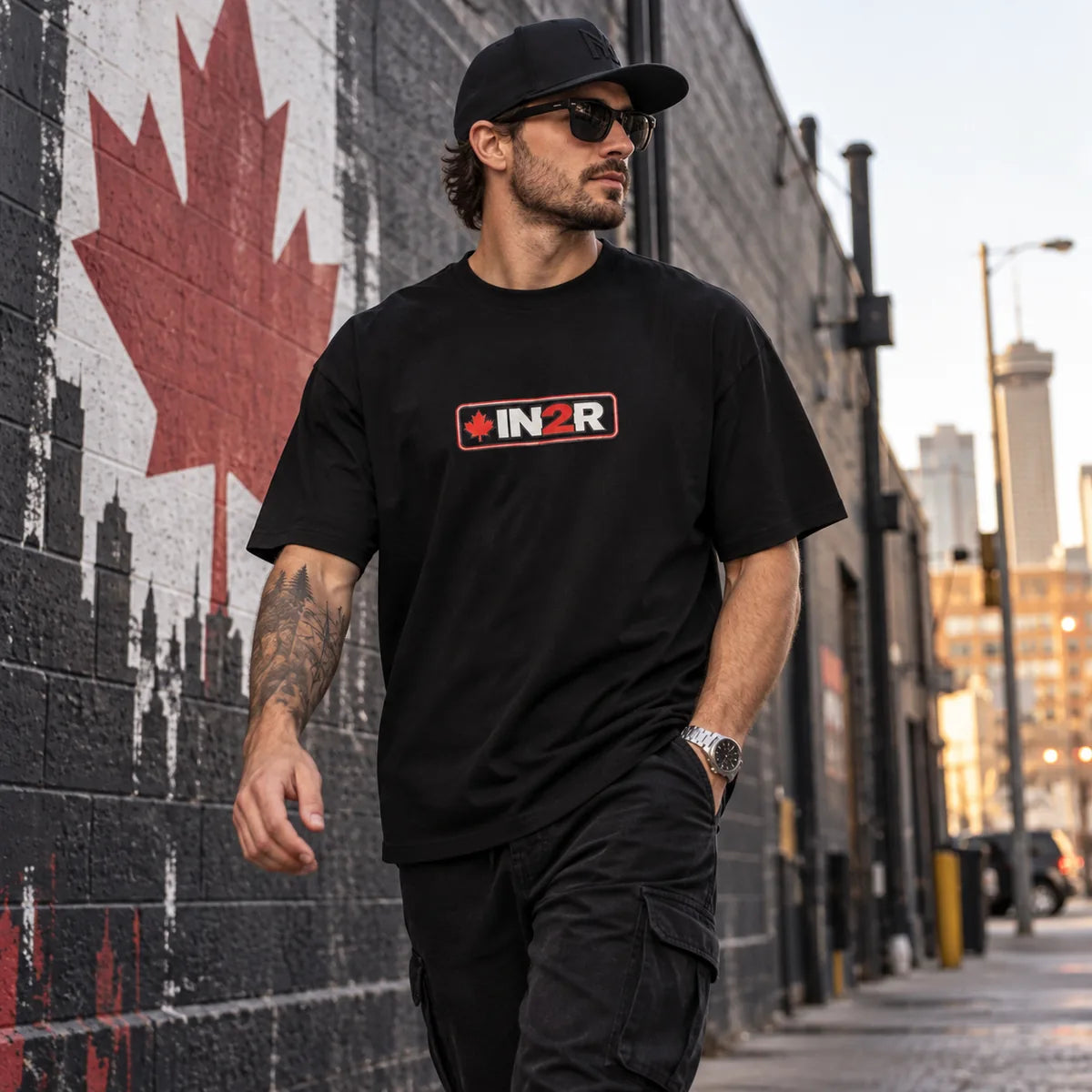 A man wearing a black t-shirt with a red and white 'IN2R' logo walks down an urban street next to a brick wall featuring a large red maple leaf mural.