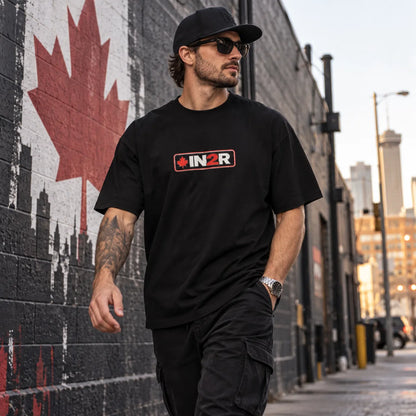 A man wearing a black t-shirt with a red and white 'IN2R' logo walks down an urban street next to a brick wall featuring a large red maple leaf mural.