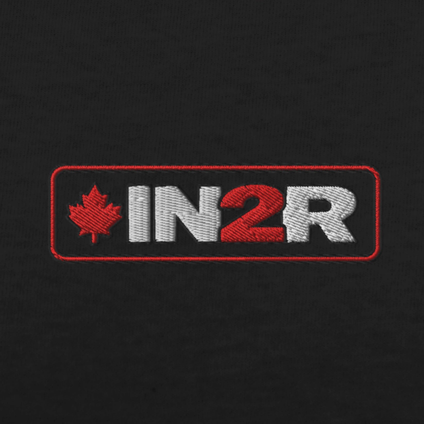 Close-up of an embroidered rectangular logo on black fabric, featuring a red maple leaf and the letters 'IN2R' in white and red.