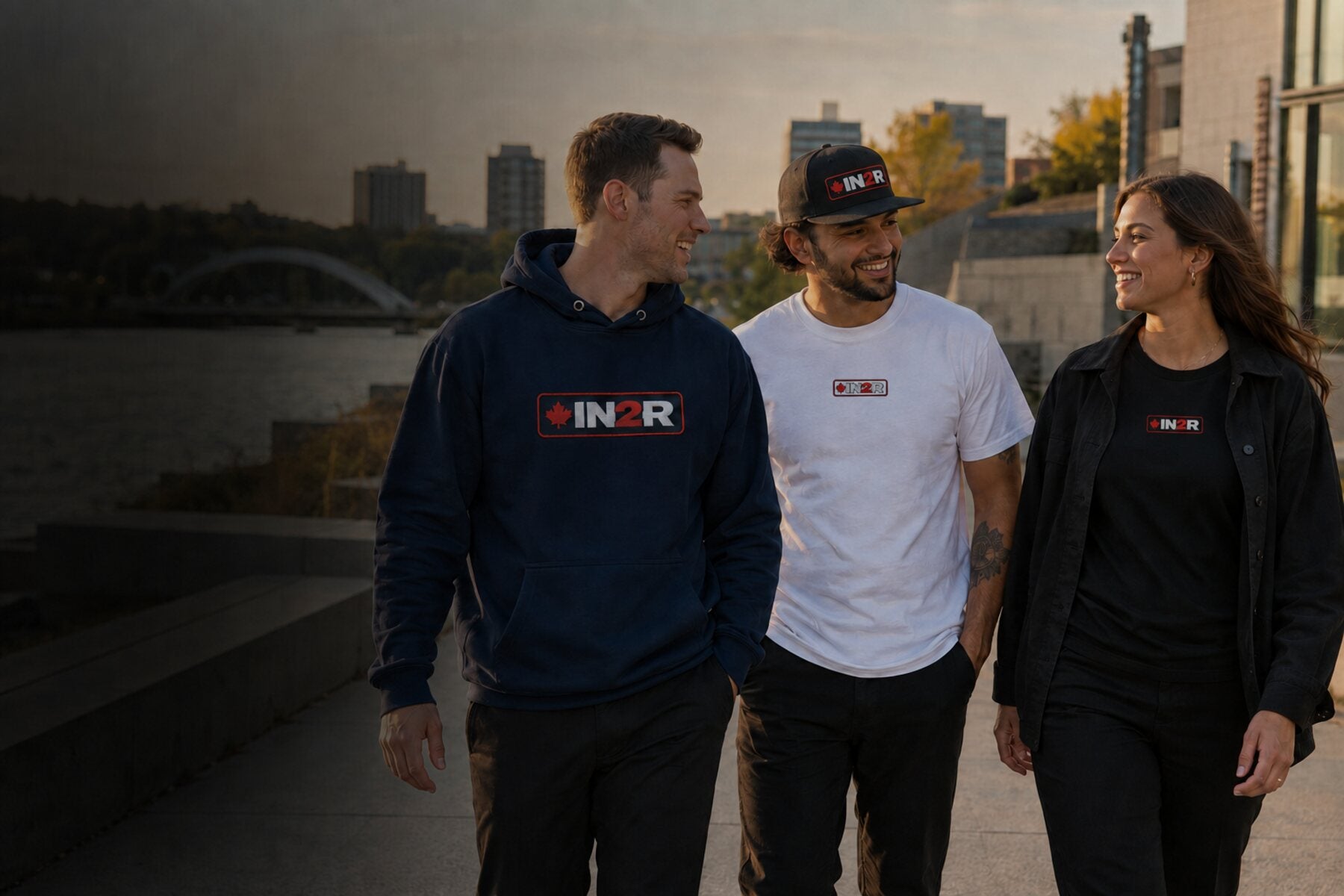 Three people walking outdoors on a waterfront promenade at sunset, each wearing a black, white, or navy hoodie or t-shirt with a matching 'IN2R' logo on the chest.