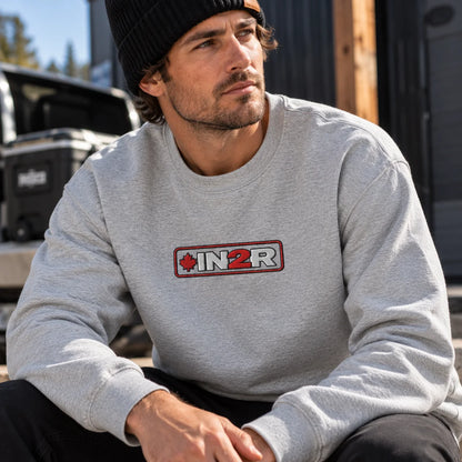 Man wearing a carbon grey IN2R Canadian Edition crewneck sweatshirt with embroidered red maple leaf logo and black knit beanie in a rugged outdoor setting.