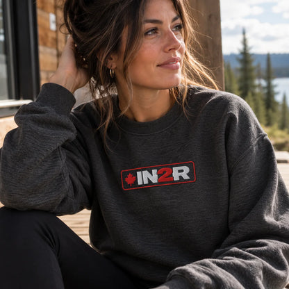 Woman wearing a charcoal heather IN2R Canadian Edition crewneck sweatshirt with red and white embroidered maple leaf logo, relaxing on a cabin porch.