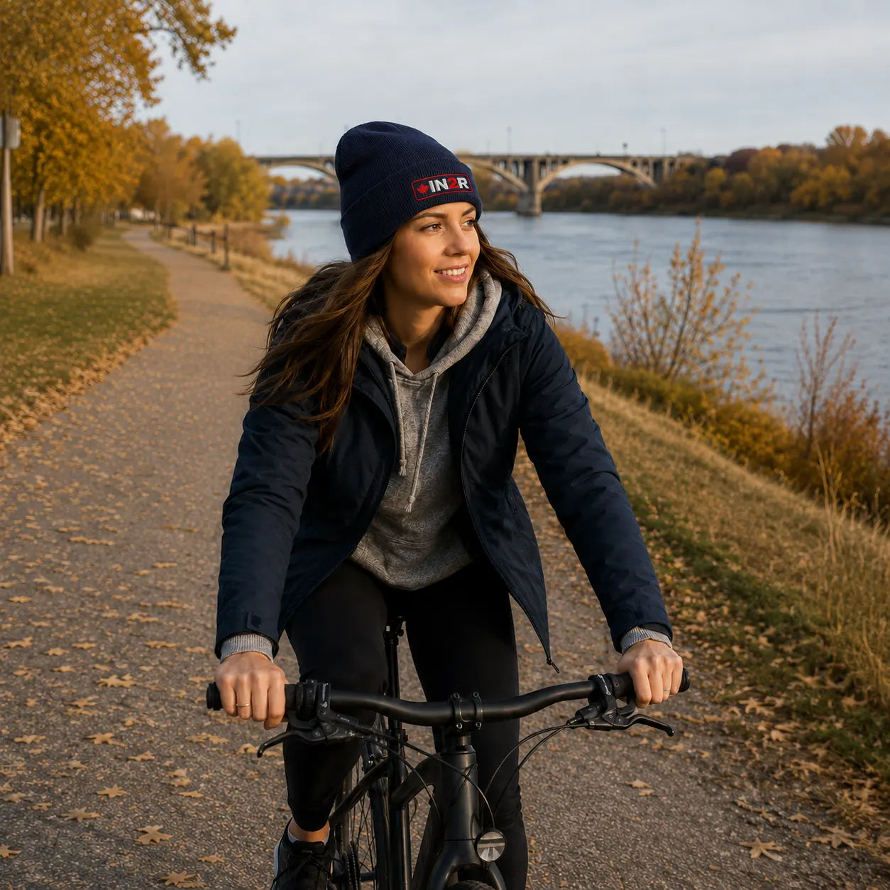 A woman riding a bicycle on an autumn path next to a river, wearing a navy blue beanie with a red and white 'IN2R' logo, a navy jacket, and a gray hoodie.