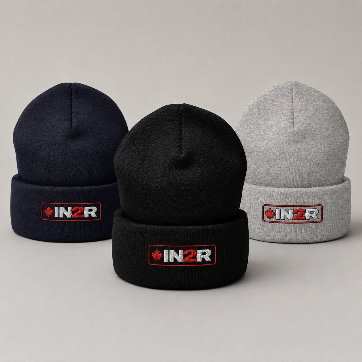 Three knit cuffed beanies on a light gray studio background: one navy, one black, and one light gray. Each beanie has a folded cuff with an embroidered IN2R logo patch in red, white, and black.