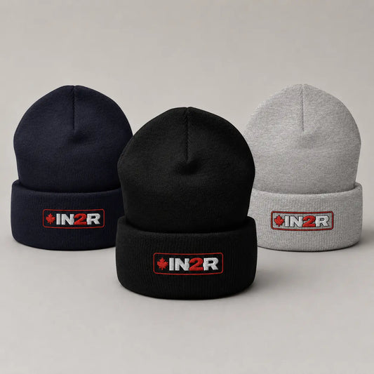 Three knit cuffed beanies on a light gray studio background: one navy, one black, and one light gray. Each beanie has a folded cuff with an embroidered IN2R logo patch in red, white, and black.