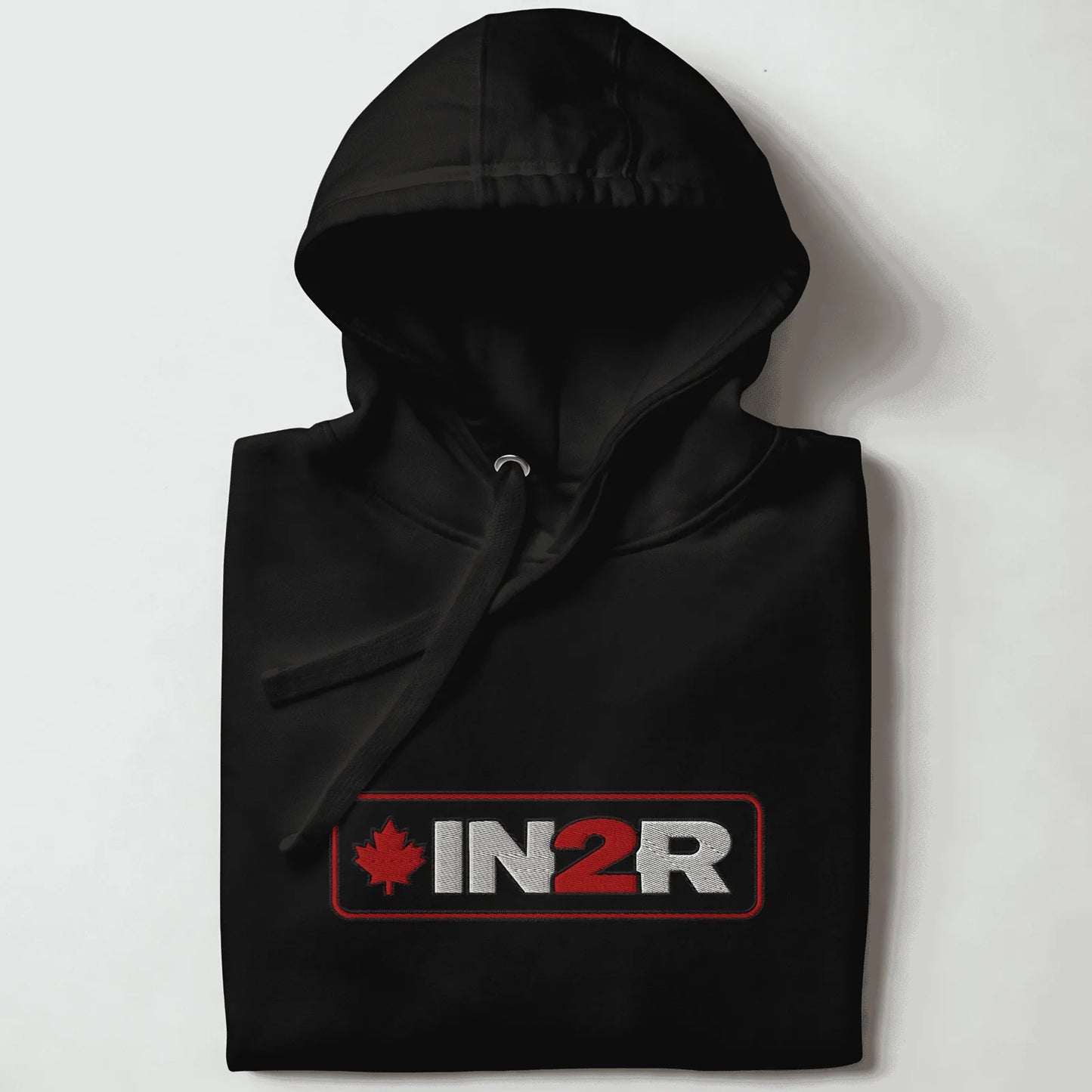 A folded black pullover hoodie with a drawstring hood, featuring a rectangular chest graphic that reads 'IN2R' in white and red with a red maple leaf on the left, set against a plain light background.