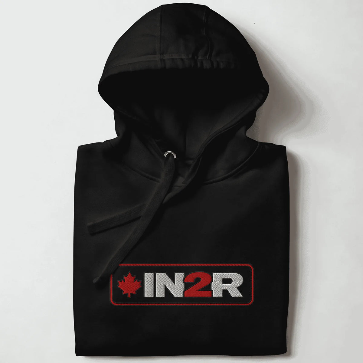 A folded black pullover hoodie with a drawstring hood, featuring a rectangular chest graphic that reads 'IN2R' in white and red with a red maple leaf on the left, set against a plain light background.
