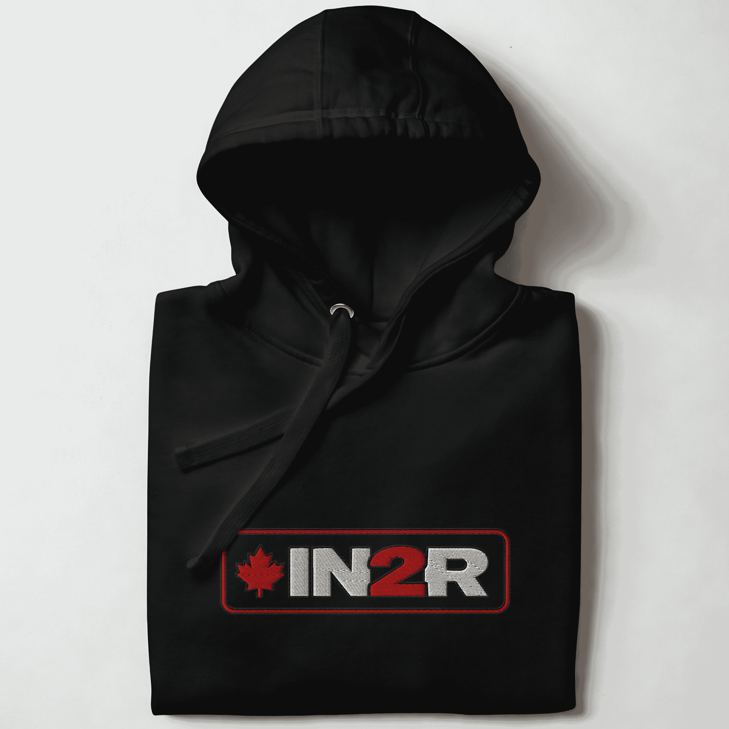 A folded black pullover hoodie with a drawstring hood, featuring a rectangular chest graphic that reads 'IN2R' in white and red with a red maple leaf on the left, set against a plain light background.