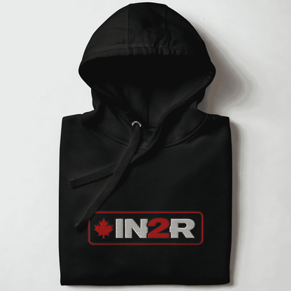 A folded black pullover hoodie with a drawstring hood, featuring a rectangular chest graphic that reads 'IN2R' in white and red with a red maple leaf on the left, set against a plain light background.