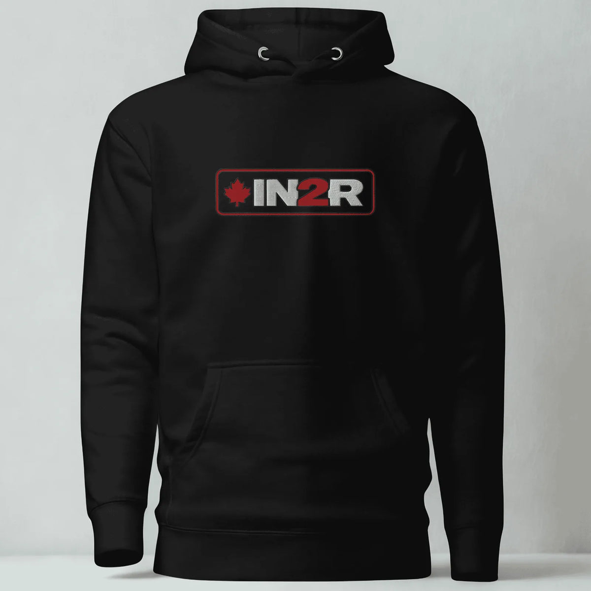 A black pullover hoodie with a front kangaroo pocket and a chest graphic that reads 'IN2R' in white and red with a small red maple leaf, shown against a light gray studio background.
