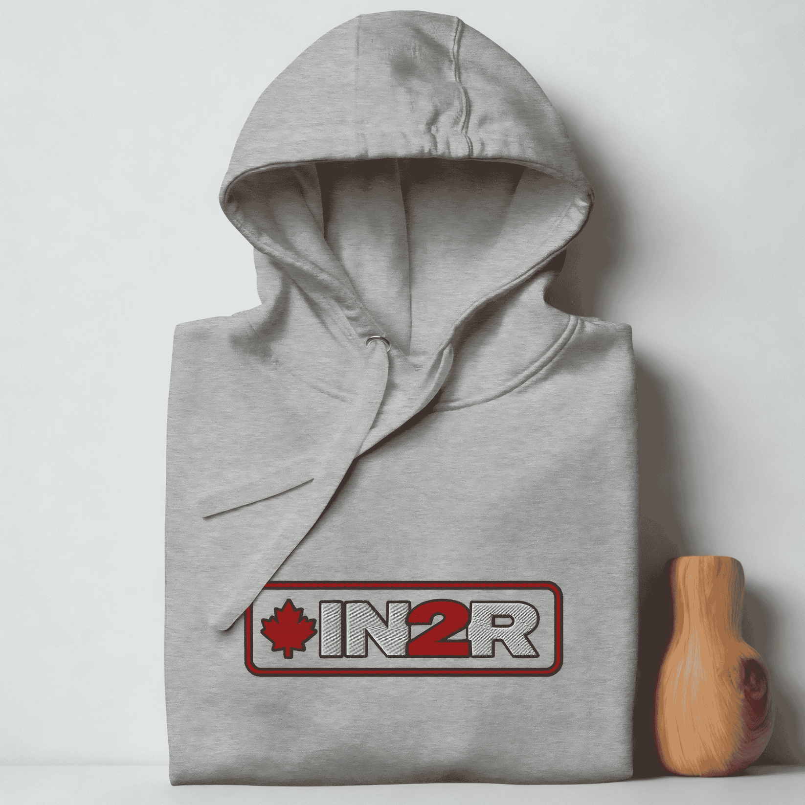 A folded gray hoodie with a drawstring hood, featuring a red and gray 'IN2R' logo with a maple leaf on the chest, set against a light gray background.