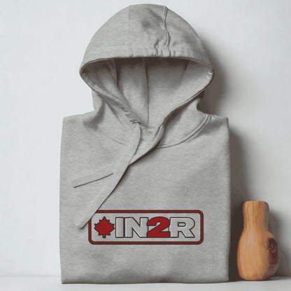 A folded light gray heather hoodie with a drawstring hood and a front kangaroo pocket, featuring a red and white 'IN2R' logo with a maple leaf on the chest. The hoodie is displayed next to a small wooden block against a plain light gray background.