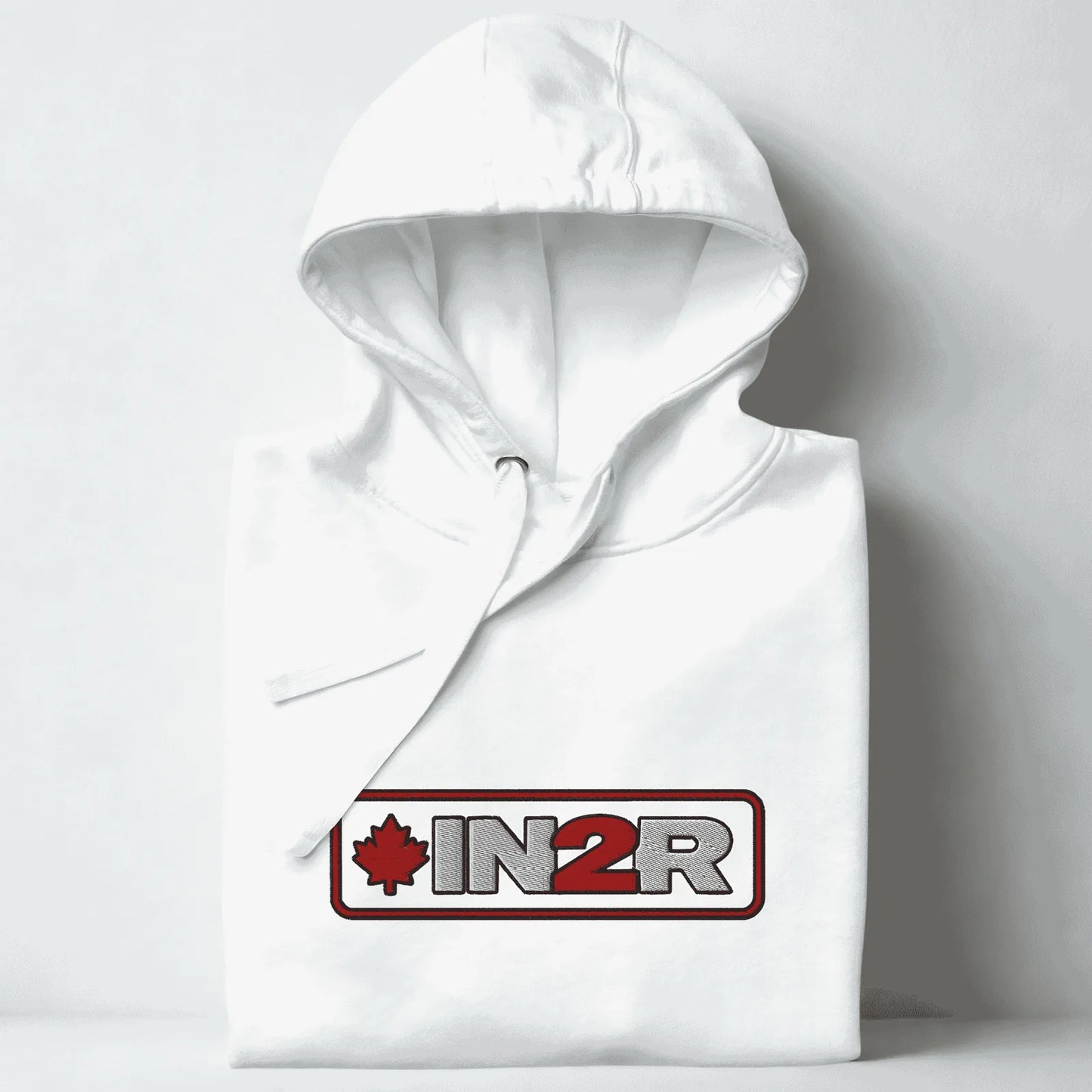 A folded white hoodie with a drawstring hood, featuring a red and gray 'IN2R' logo with a maple leaf on the chest, set against a light gray background.