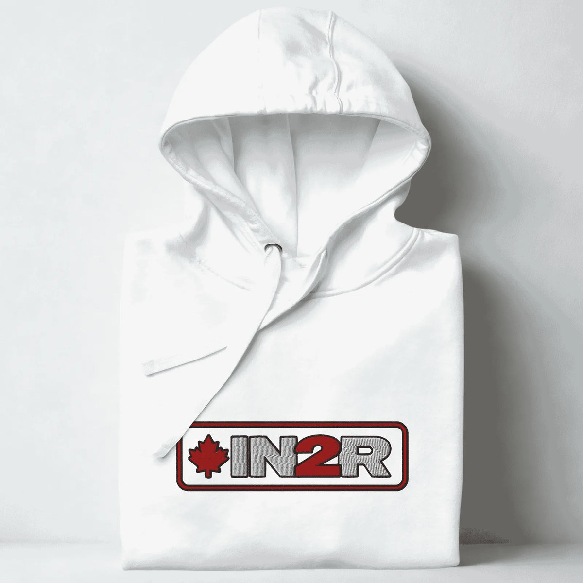 A folded white hoodie with a drawstring hood, featuring a red and gray 'IN2R' logo with a maple leaf on the chest, set against a light gray background.