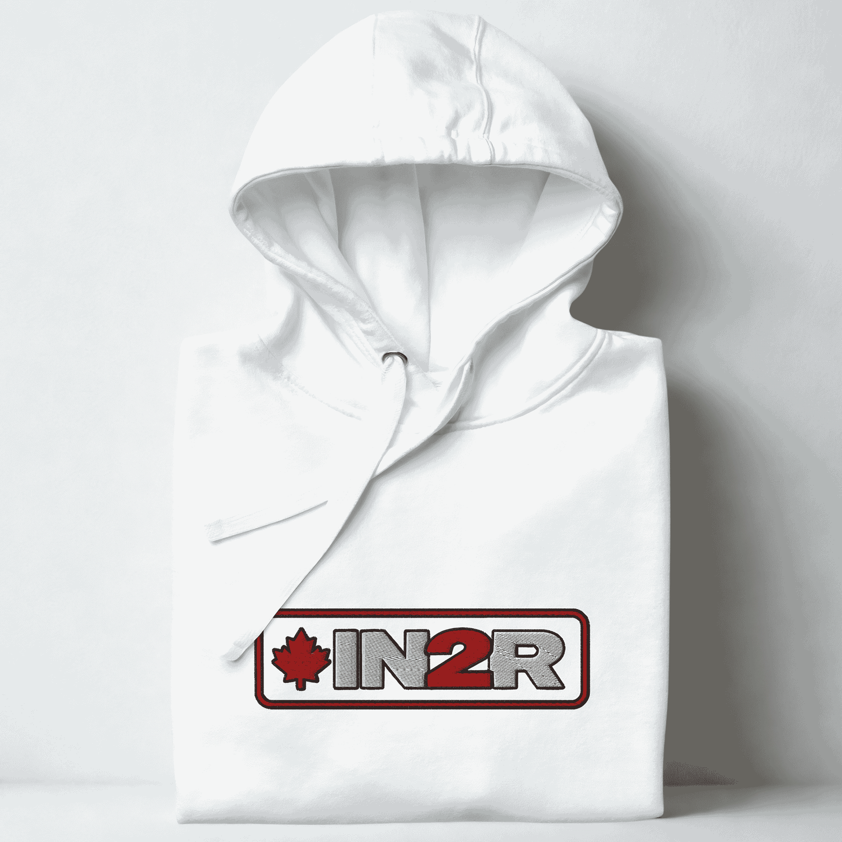 A folded white hoodie with a drawstring hood, featuring a red and gray 'IN2R' logo with a maple leaf on the chest, set against a light gray background.