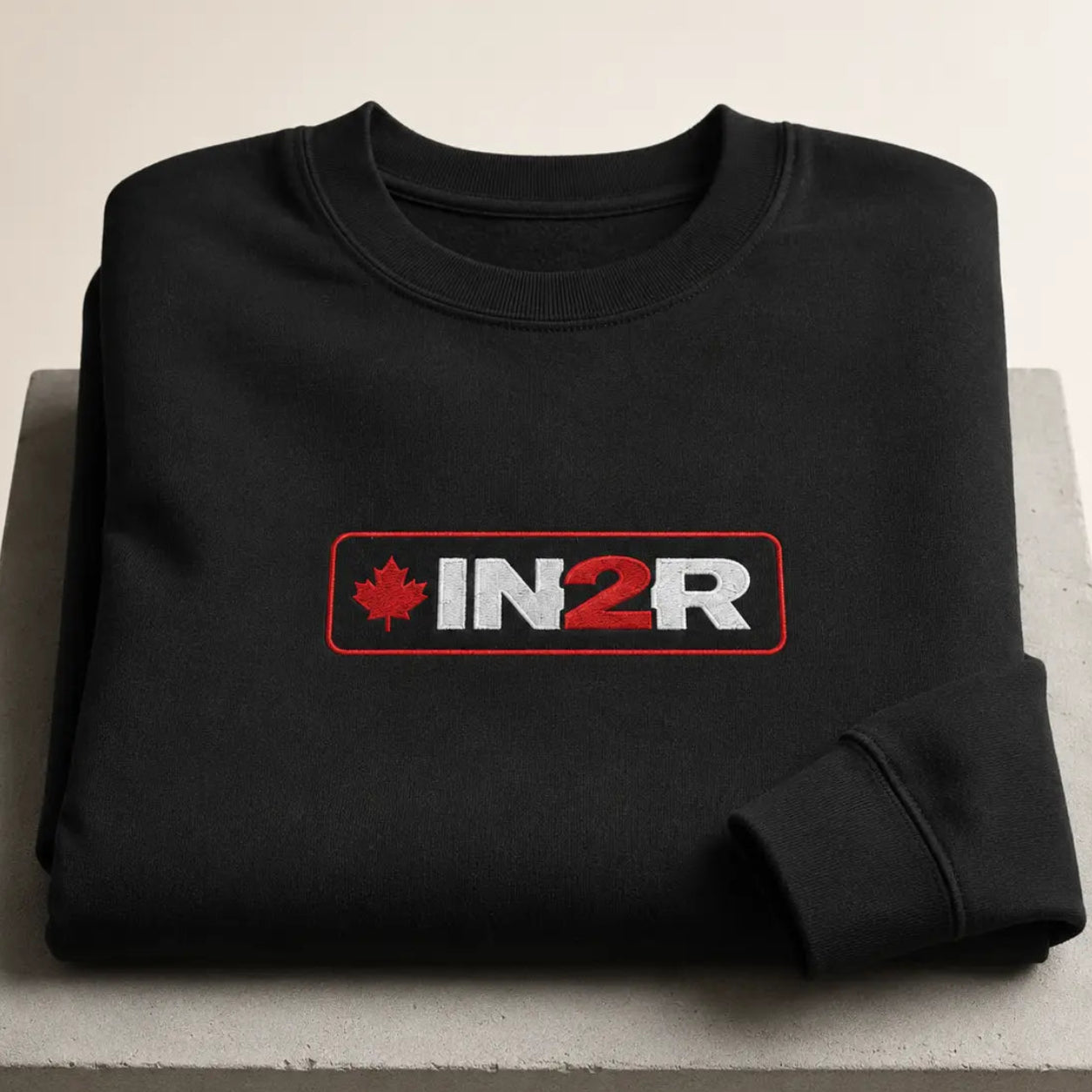 A folded black crewneck sweatshirt displayed on a light gray concrete block. The sweatshirt features a rectangular chest graphic with a red maple leaf, the text 'IN2R' in white and red, and a red border.
