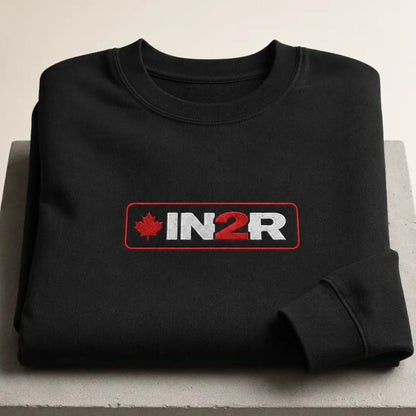A folded black crewneck sweatshirt displayed on a light gray concrete block. The sweatshirt features a rectangular chest graphic with a red maple leaf, the text 'IN2R' in white and red, and a red border.