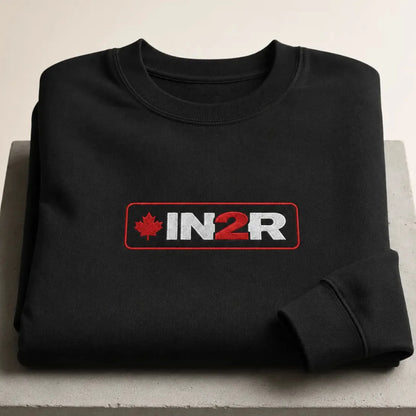A folded black crewneck sweatshirt with a red and white 'IN2R' logo and a red maple leaf on the chest, displayed on a light gray surface.
