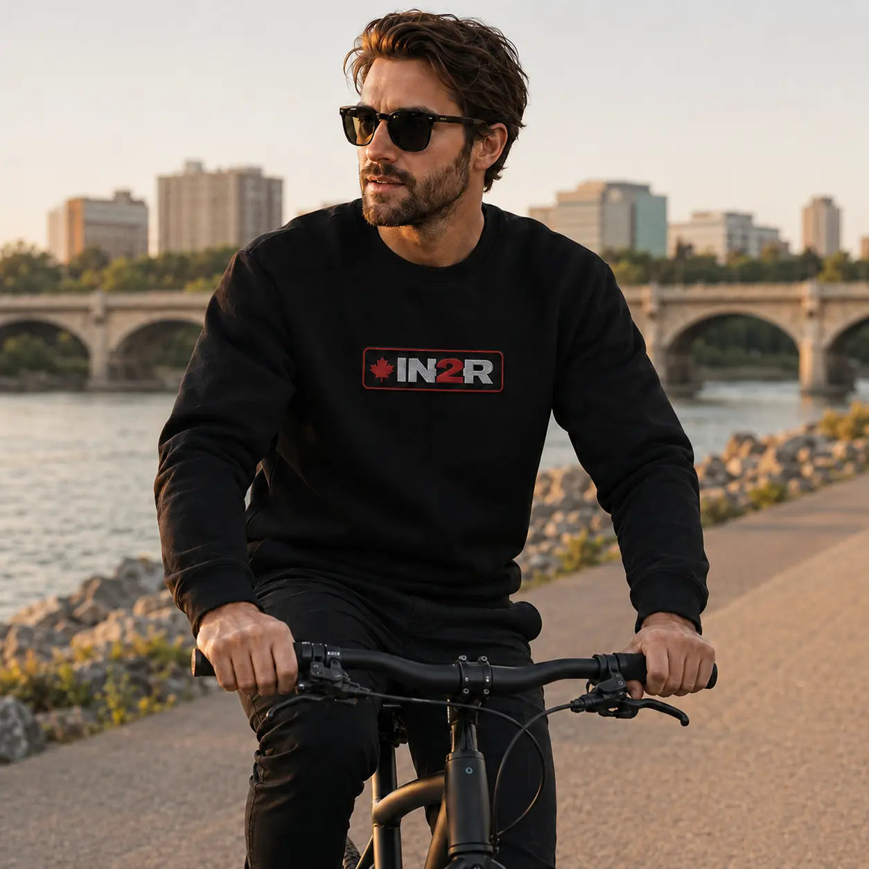 A man wearing a black crewneck sweatshirt with a red and white 'IN2R' logo on the chest, riding a black bicycle on a paved path next to a river with a city skyline in the background.