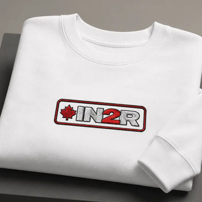A folded white crewneck sweatshirt displayed on a dark gray block, featuring a rectangular chest graphic with a red maple leaf, the text 'IN2R' in white and red, and a red border.
