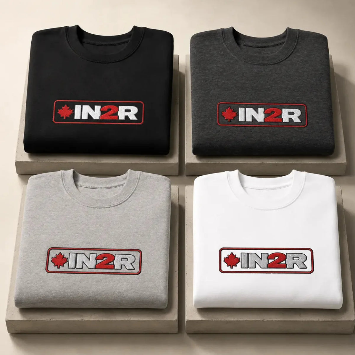 Four folded crewneck t-shirts in black, dark gray, light gray, and white, arranged in a 2x2 grid on beige display blocks. Each shirt features a rectangular chest logo with a red maple leaf, the text 'IN2R' in white, and a red border.