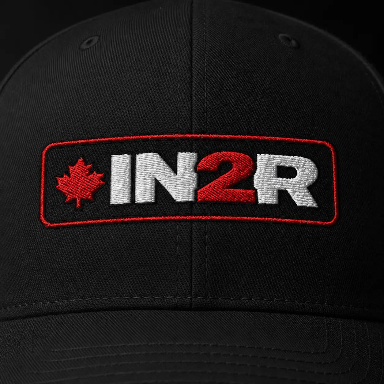 Close-up of a black baseball cap featuring an embroidered rectangular patch with a red maple leaf and the text 'IN2R' in white and red.