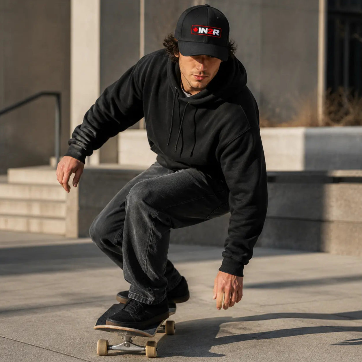 A man in a black hoodie and black cap with a red and white 'IN2R' logo is crouching on a skateboard in an outdoor concrete plaza.