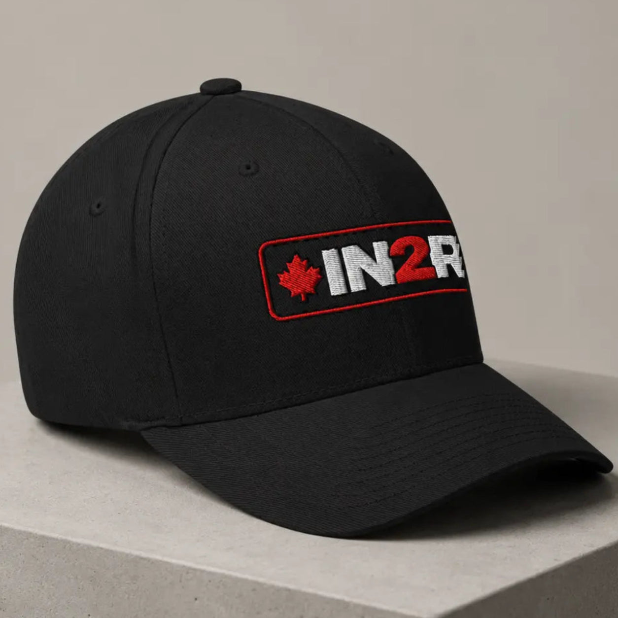 A black baseball cap with a curved brim, featuring a front patch that reads 'IN2R' in white and red with a red maple leaf, displayed on a light gray concrete pedestal against a neutral beige background.