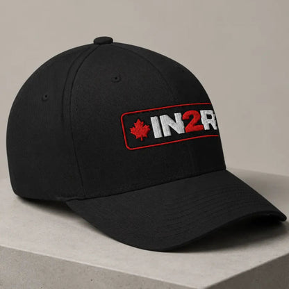 A black baseball cap with a curved brim, featuring a front patch that reads 'IN2R' in white and red with a red maple leaf, displayed on a light gray concrete pedestal against a neutral beige background.