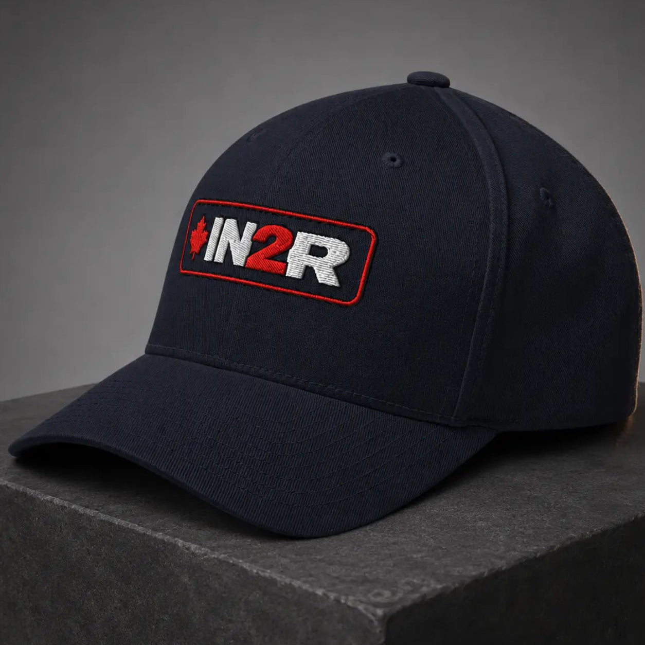 A dark navy blue baseball cap with a curved brim, featuring a rectangular embroidered patch on the front with the text 'IN2R' in white and red with a small red maple leaf. The cap is displayed on a dark gray textured block against a dark gray gradient background.
