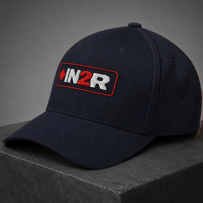 A dark navy blue baseball cap with a rectangular embroidered patch on the front that reads 'IN2R' in white and red with a small red maple leaf, resting on a dark gray stone block against a gray background.
