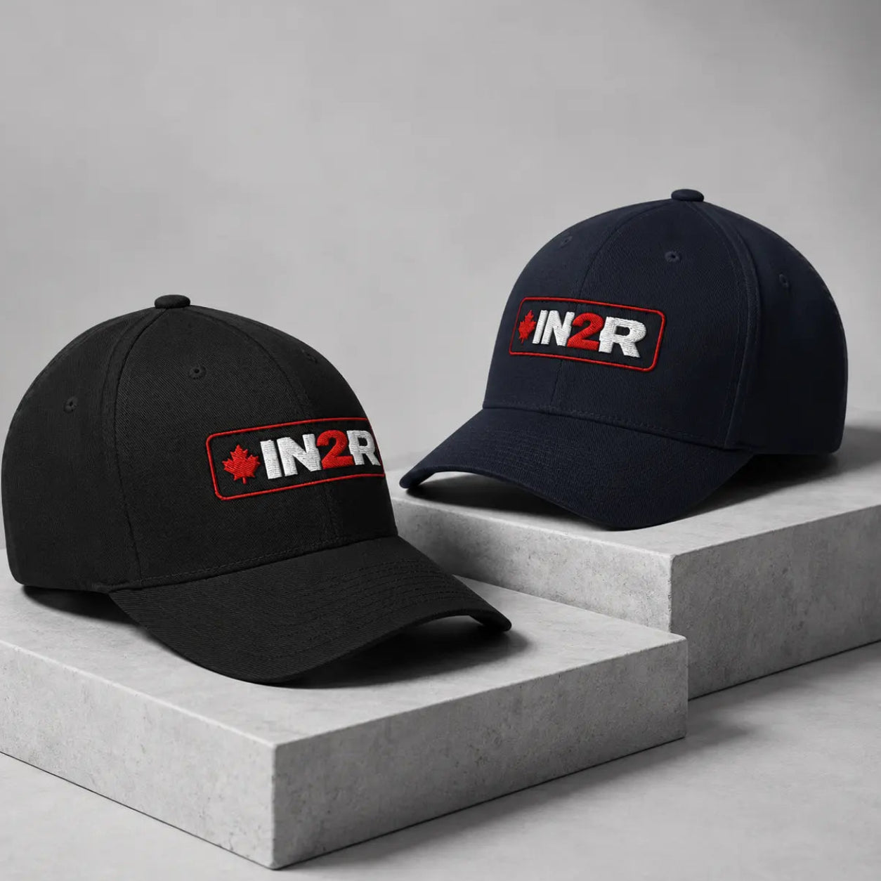 Two baseball caps displayed on concrete blocks against a gray studio background. The left cap is black with a red and white embroidered logo, and the right cap is navy blue with a matching red and white embroidered logo.