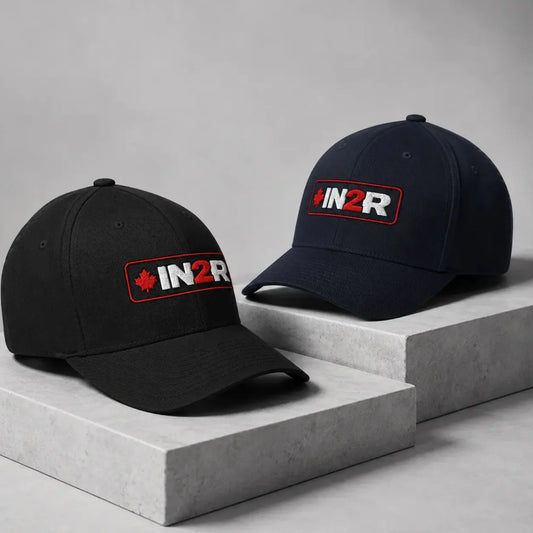 Two baseball caps displayed on concrete blocks against a gray studio background. One cap is black and the other is navy blue, both featuring a red and white 'IN2R' logo patch on the front.
