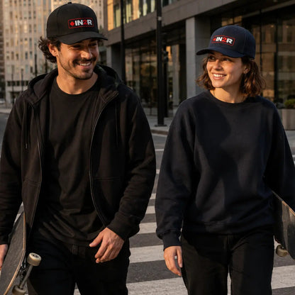 A man and a woman walking on a city crosswalk, both wearing black IN2R brand caps, black tops, and black pants. The man holds a skateboard, and the woman holds a phone.