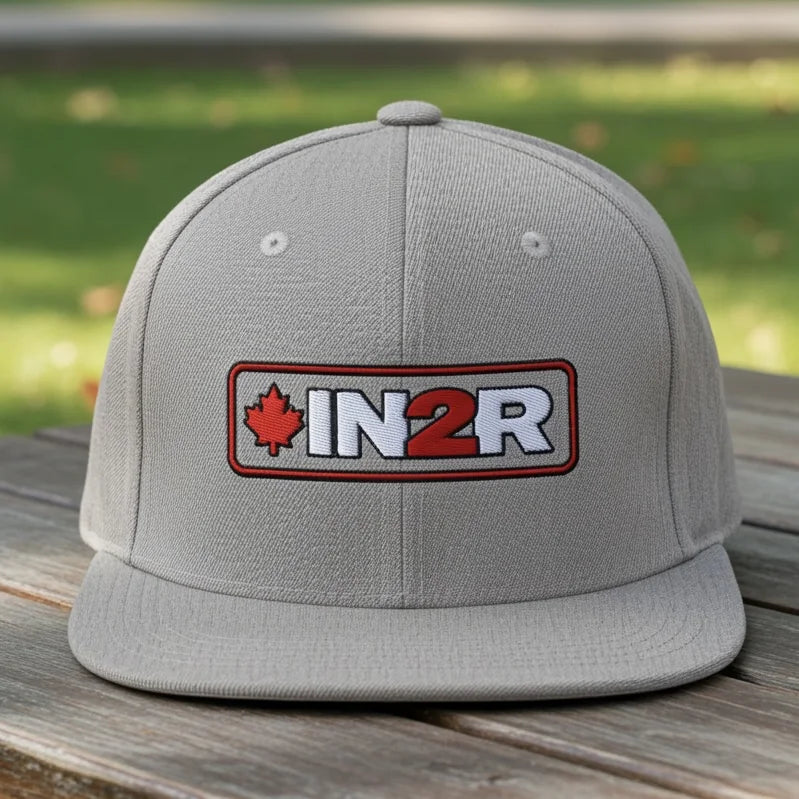 IN2R Canadian Edition grey snapback hat with flat brim and embroidered red maple leaf, white IN2R lettering with red 2, displayed on a wooden table outdoors.