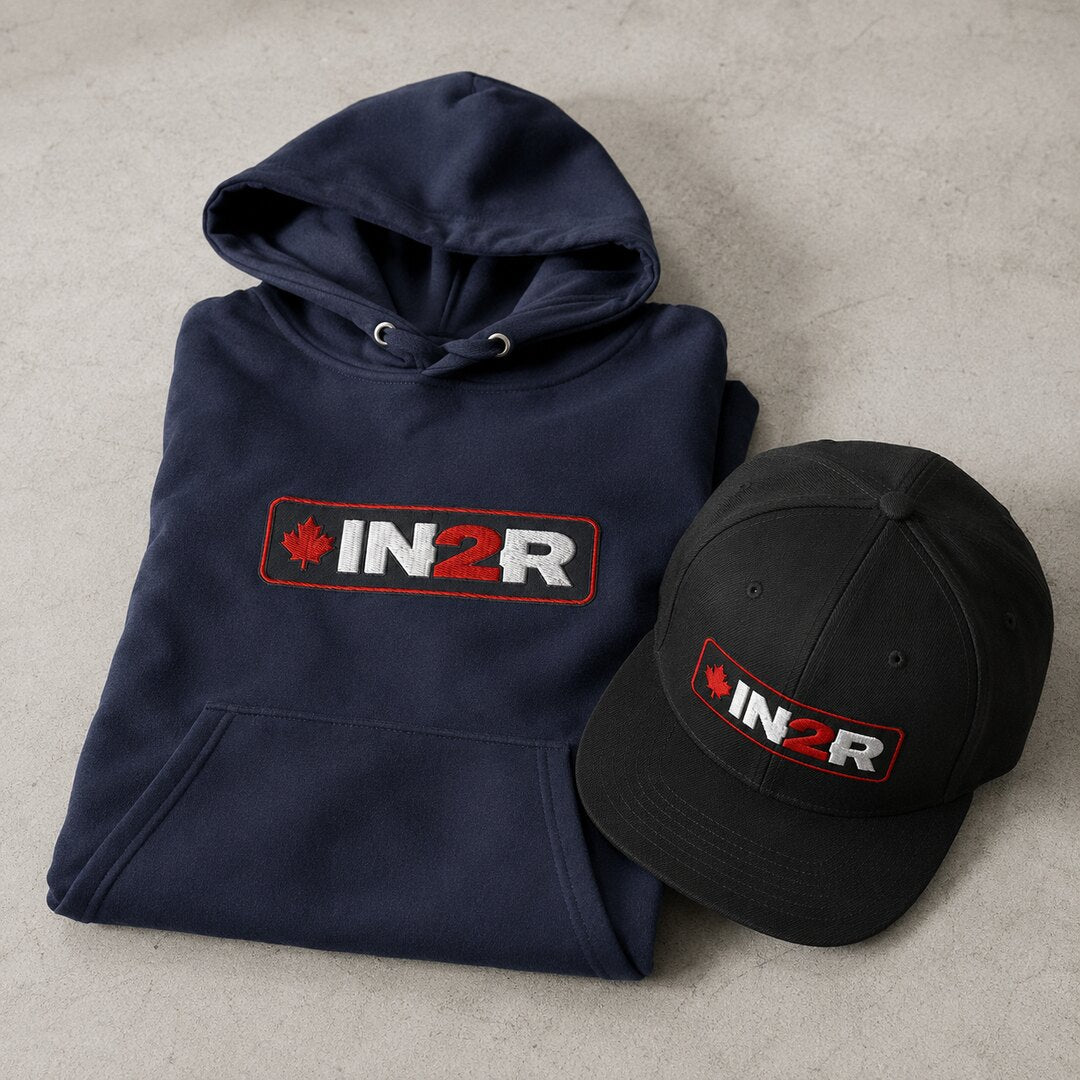 A navy blue hoodie and a black baseball cap, both folded and arranged on a light gray textured surface. Each item features an embroidered 'IN2R' logo with a red maple leaf on the front.