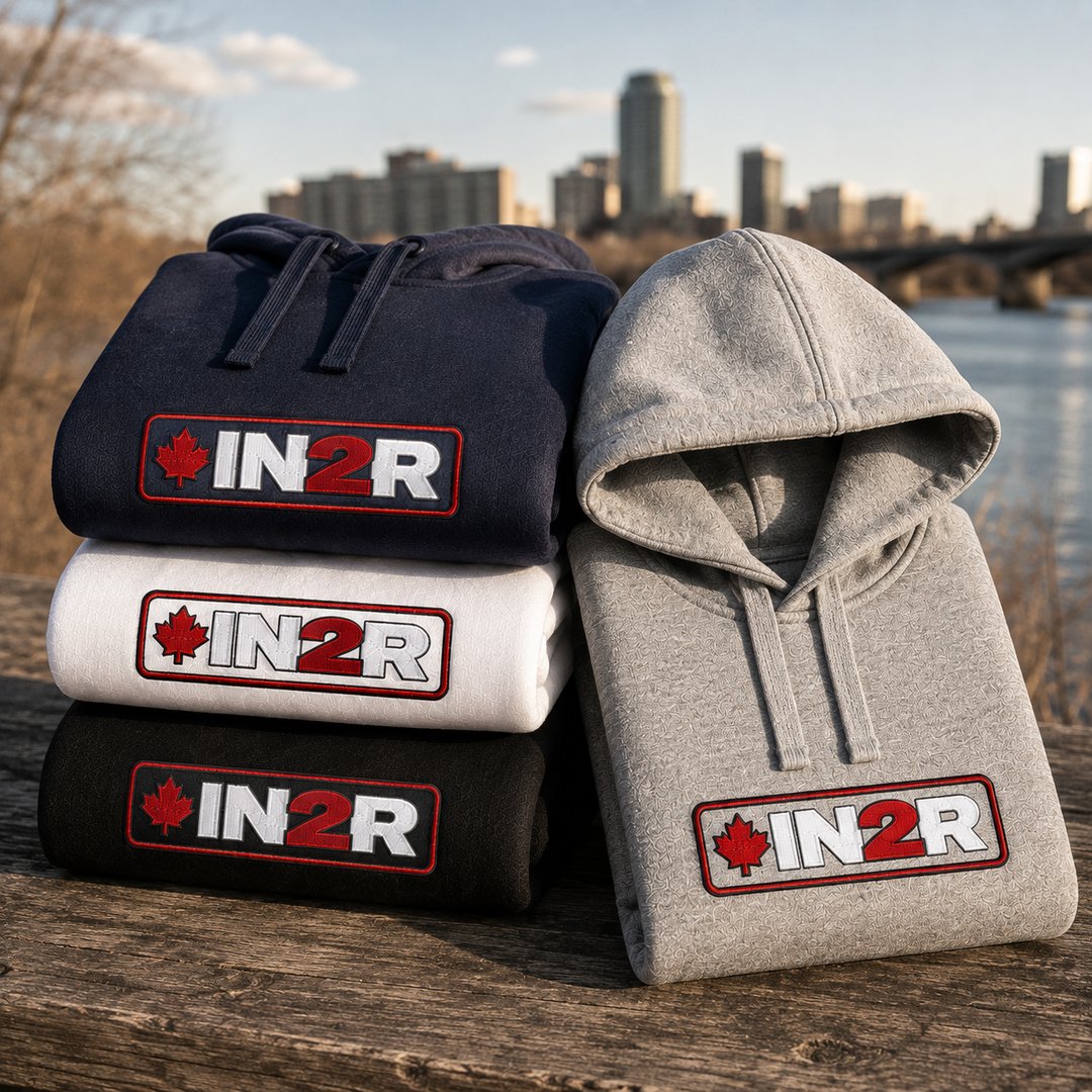 IN2R Canadian Edition hoodies in navy, white, black and carbon grey stacked outdoors beside a riverfront, featuring embroidered red, white and black IN2R maple leaf logos.