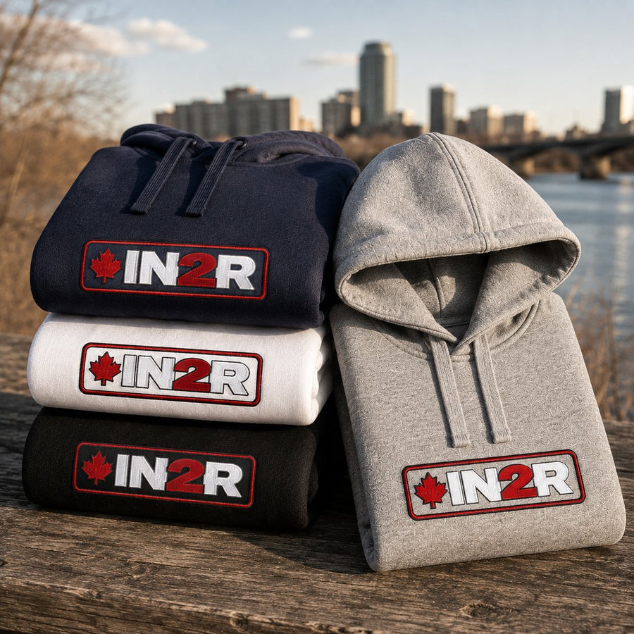 IN2R Canadian Edition hoodies in navy, white, black and carbon grey stacked outdoors beside a riverfront, featuring embroidered red, white and black IN2R maple leaf logos.