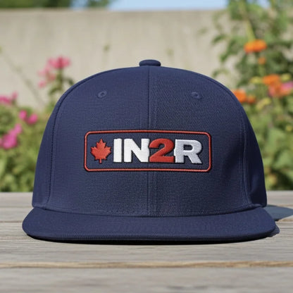IN2R Canadian Edition navy blue snapback hat with flat brim, red maple leaf and white and red embroidered IN2R logo, photographed outdoors on a wooden table with flowers in the background.