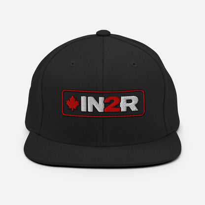 A black snapback cap featuring a rectangular embroidered patch on the front with a red maple leaf, the text 'IN2R' in white and red, and a red border.
