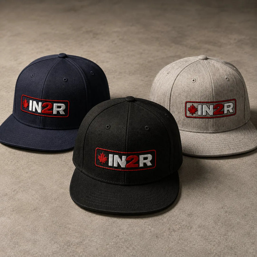 Three flat-brim snapback caps arranged on a textured neutral surface: one navy, one black, and one light gray. Each cap has an embroidered front patch reading IN2R with a small red maple leaf.