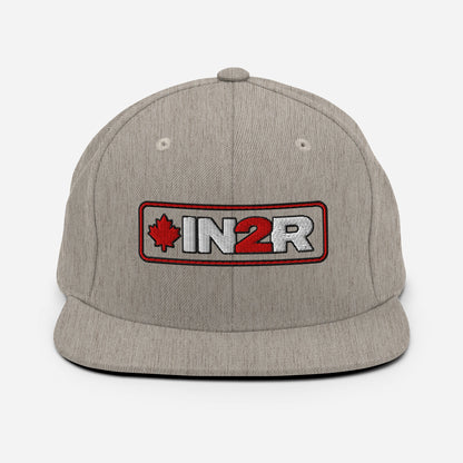 A heather gray snapback cap featuring a rectangular embroidered patch on the front with a red maple leaf, the text 'IN2R' in white and red, and a red border.