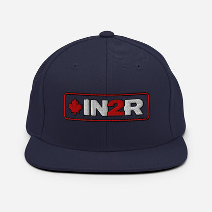 A black snapback cap featuring a rectangular embroidered patch on the front with a red maple leaf, the text 'IN2R' in white and red, and a red border.
