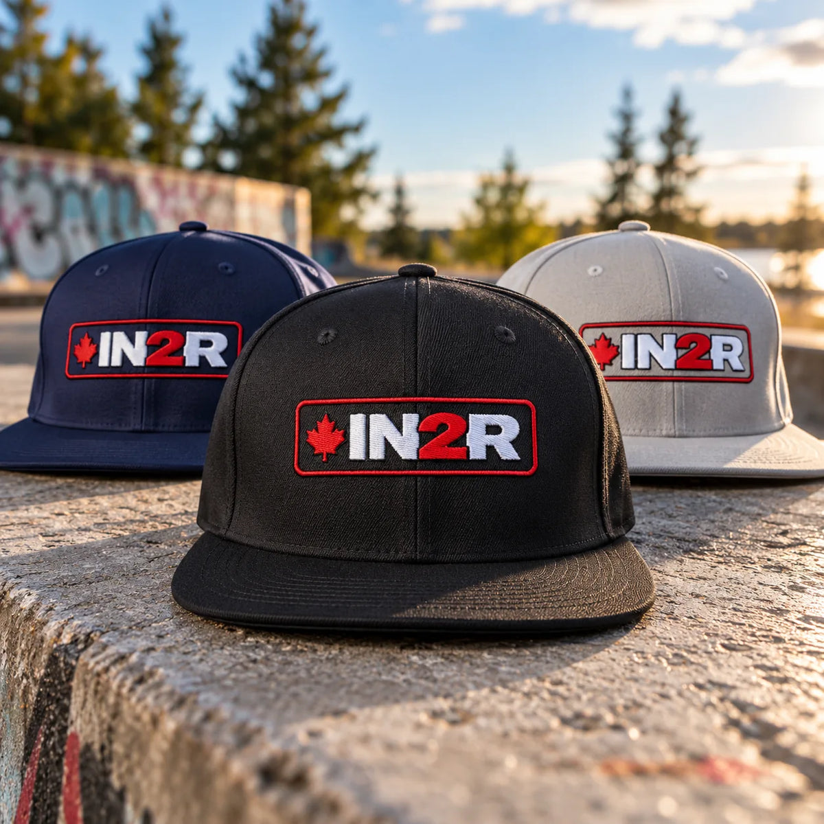 IN2R Canadian Edition snapback hat collection in black, navy blue and grey, featuring embroidered red maple leaf and red and white IN2R logos, displayed outdoors on concrete in warm sunlight.